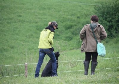 Educatrice in giacca gialla lavora con un cane nero seduto accanto a una proprietaria